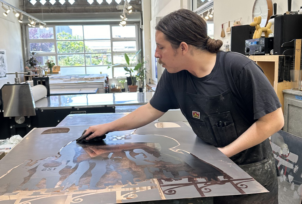 A printmaker in a black apron wipes a large metal printing plate on a worktable in a bright printmaking studio.